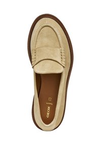 Mocasines de ante beige con una suela de goma marrón plana, que presentan un diseño de mocasín cosido y un estilo deslizante. Detalles del logo en dorado en el interior.