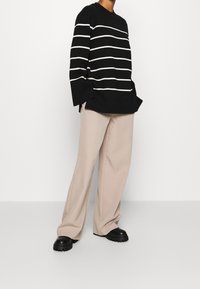 Maglione a righe bianche e nere con maniche larghe abbinato a pantaloni beige a gamba larga e scarpe con suola spessa nere. Texture lisce in tutto l'outfit.