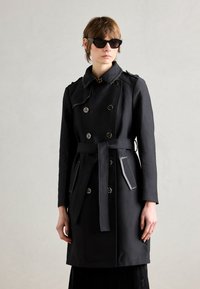 Manteau noir à double boutonnage, avec épaulettes, taille ceinturée et garniture des poches contrastante. Porté avec des lunettes de soleil noires angulaires.