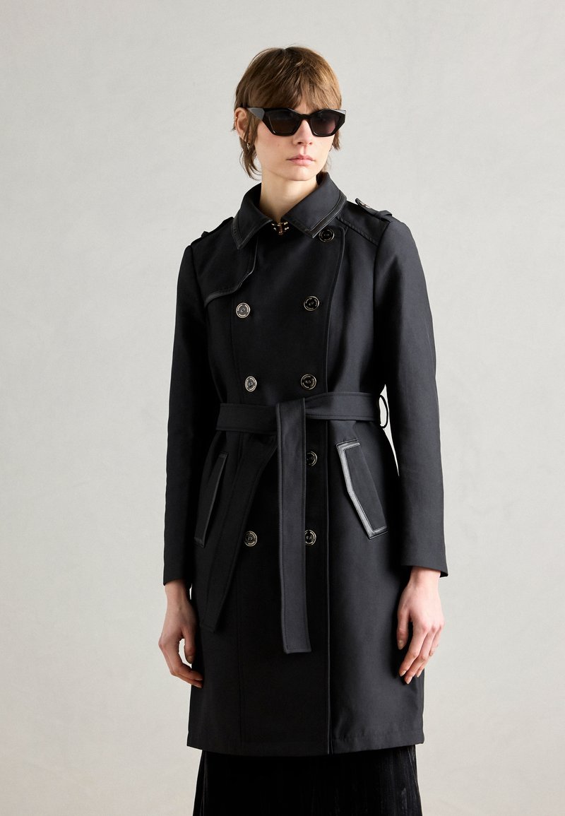 Manteau noir à double boutonnage, avec épaulettes, taille ceinturée et garniture des poches contrastante. Porté avec des lunettes de soleil noires angulaires.
