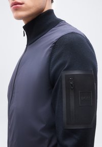 Άνδρας που φοράει ναυτικό μπουφάν με ψηλό γιακά, με μαύρη τσέπη με φερμουάρ στην αριστερή πάνω πλευρά του μπράτσου, με την ετικέτα "BOSS", εμφανιζόμενος από το λαιμό έως το μέσο του κορμού.