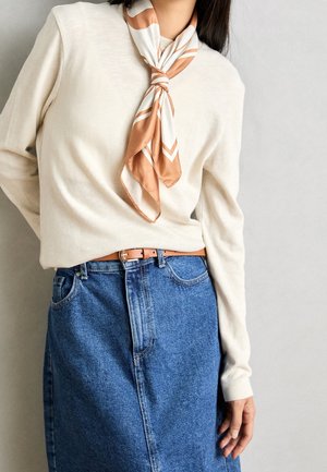 Osoba ubrana w kremowy sweter, niebieską dżinsową spódnicę z brązowym paskiem oraz beżowo-biały paskowany szalik zawiązany wokół szyi, na jednolitym tle.