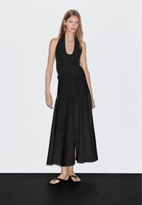 Abito nero con scollo a halter e gonna fluida, di lunghezza fino al ginocchio, con spacchi laterali, realizzato in tessuto leggero, abbinato a sandali neri piatti.