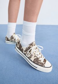 Baskets en toile à motifs marron et crème avec des lacets blancs, portées avec des chaussettes blanches côtelées, posées sur une surface bleu clair.
