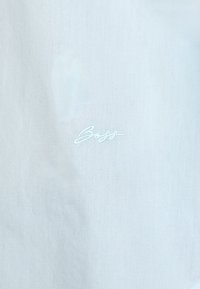 Lyseblåt stoff med en glatt tekstur har et brodert logo i en litt mørkere nyanse. Designet er minimalistisk og moderne.