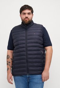 Kara zila pufīga veste ar augstu apkakli, priekšējo rāvējslēdzēju, sānu kabatām un horizontālu quiltēta dizaina. Valkā uz kara zila īsām piedurknēm krekla.