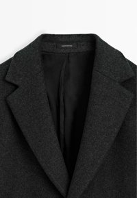 Cappotto di lana grigio scuro con ampi revers, apertura frontale e fodera interna nera, etichetta "Made in Portugal" sul colletto.