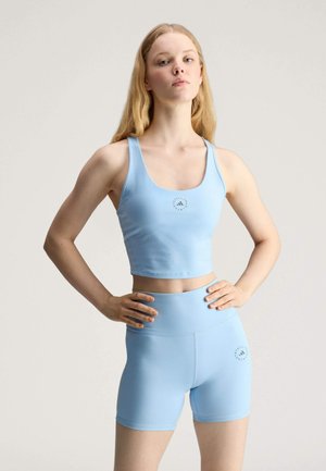 Junge Frau steht mit den Händen in der Hüfte, trägt ein hellblaues sportliches Croptop und passende hoch taillierte Shorts vor einem schlichten Hintergrund.