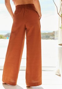 Pantalons larges en orange brûlé, fabriqués en tissu léger. Taille élastique et poches latérales, avec une texture lisse et un subtil éclat.