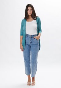 Cardigan turquesa, de manga corta, con frente abierto. Top blanco sin mangas, jeans de tiro alto en azul y sandalias blancas con tiras. Tejido suave y corte relajado.