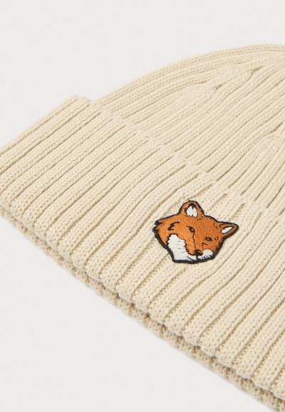 Knit beanie in light beige with ribbed texture. Features an embroidered fox face patch in orange and white, positioned on the front.