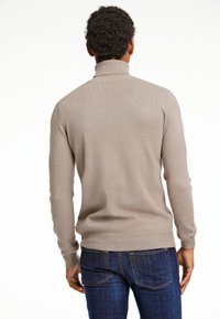 Beiger Pullover mit hohem Kragen, geripptem Textur und tailliertem Schnitt. Getragen mit dunkelblauen Jeans, die einen geraden Schnitt und Gesäßtaschen aufweisen.