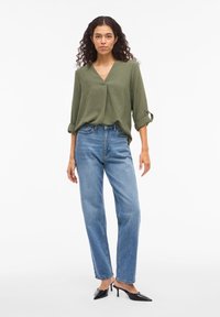 Λαδί πράσινη μπλούζα με V λαιμόκοψη, μανίκια γυρισμένα· συνδυασμένη με ανοιχτό μπλε ψηλόμεσα τζιν παντελόνια με ίσια γραμμή και μαύρα ψηλοτάκουνα παπούτσια με μυτερή μύτη.