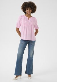 Rozā blūze ar V veida kaklu un dekoratīvām šuvēm, savienota ar gaiši ziliem izvērstiem džinsiem un baltiem smailiem papēžiem. Strukturēts audums rada mīkstu izskatu.