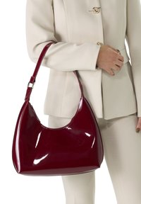 Sac hobo en cuir verni bordeaux avec une silhouette courbée, une poignée unique et une finition brillante. Dispose d'une couture le long du bord supérieur.