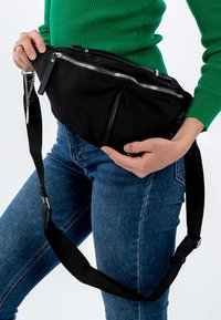 Černá látková ledvinka s několika kapsami na zip, nastavitelným popruhem a kovovým kováním. Má elegantní, kompaktní design.