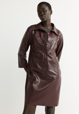 Woman wearing a dark brown leather button-up dress with long sleeves and pockets over a gray top, standing against a plain light background.