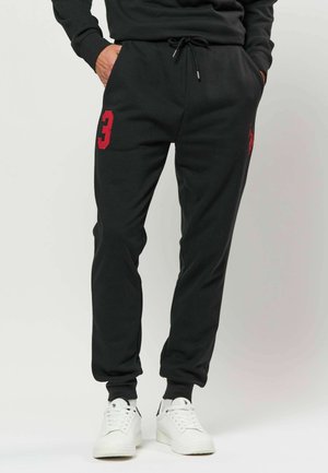 Personne portant un pantalon de jogging noir avec le chiffre trois rouge sur la cuisse gauche et des baskets blanches, mains dans les poches.