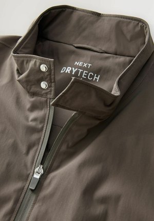Bruine waterdichte jas met een staande kraag, ritssluiting en drukknoopsluiting. "NEXT DRYTECH" logo gedrukt aan de binnenkant van de kraag.