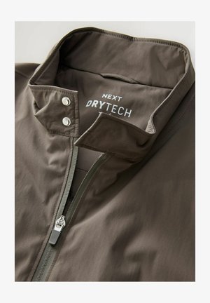 Bruine waterdichte jas met een staande kraag, ritssluiting en drukknoopsluiting. "NEXT DRYTECH" logo gedrukt aan de binnenkant van de kraag.