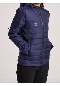 Chaqueta acolchada azul marino con capucha, que presenta acolchado horizontal, cierre de cremallera, bolsillos laterales y un logo en el pecho. Tejido suave y texturizado.