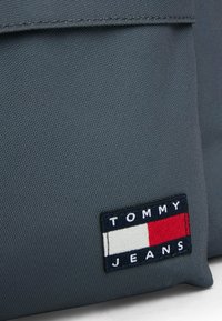 Tessuto grigio, texture liscia, con un pannello rettangolare che riporta "TOMMY JEANS" in bianco su strisce blu, rosse e bianche.