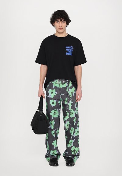 Schwarzes T-Shirt mit blauer Schrift, kombiniert mit grauen Hosen mit grünen Blumenmustern. Das Model trägt eine schwarze Tasche und schwarze Schuhe.