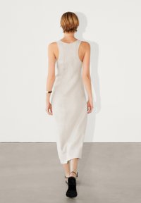 Robe midi sans manches beige clair avec une silhouette ajustée, une texture lisse et un ourlet arrondi. Des chaussures noires à bride cheville sont portées.