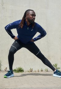 Marineblauwe lange mouw ritshemd met reflecterende accenten, gecombineerd met zwarte leggings met witte strepen, en blauwe hardloopschoenen met groene accenten.