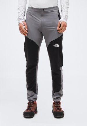 Άτομο που φορά γκρι και μαύρο παντελόνι πεζοπορίας The North Face και καφέ μπότες πεζοπορίας, στέκεται με τα χέρια κοντά στις τσέπες.