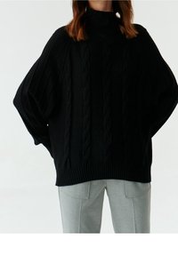 Pull à col roulé en tricot noir avec des manches amples et un ourlet côtelé, associé à un pantalon gris clair ajusté. Texture douce et coupe décontractée.