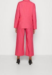 Costume rose sur mesure avec une veste légèrement oversize et un pantalon à jambes larges. Tissu lisse avec une texture soyeuse. Chaussures à talons noires complètent la tenue.