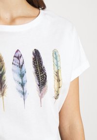 T-shirt blanc montrant quatre grandes illustrations détaillées de plumes dans des nuances de bleu, violet, noir et beige sur la zone de la poitrine.