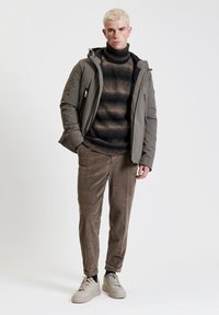Giacca imbottita verde oliva con zip, dolcevita marrone a trama, pantaloni di velluto a coste marroni e sneakers grigio chiaro. Texture e colori ricchi.