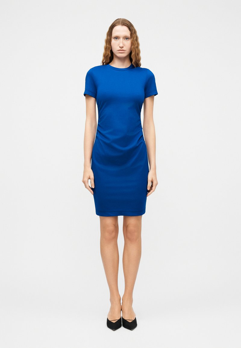 Γυναίκα με κυματιστά μαλλιά, ντυμένη με φόρεμα ριχτό μέχρι το γόνατο σε βασιλικό μπλε χρώμα και μαύρες μυτερές γόβες, που στέκεται μπροστά σε λευκό απλό φόντο.