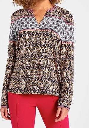 Woman wearing a long-sleeve, patterned blouse with paisley and geometric designs, paired with bright pink pants, standing against a plain background.