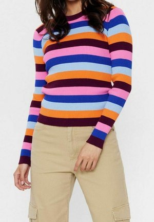Femme portant un pull à manches longues côtelé avec des rayures horizontales roses, bleues, orange et bordeaux, associé à un pantalon cargo beige.