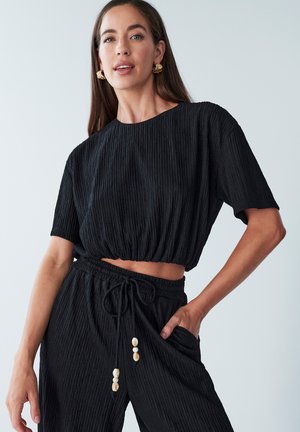 Top corto nero con texture a pieghe e maniche corte, abbinato a pantaloni neri coordinati con cordoncino e perline decorative.