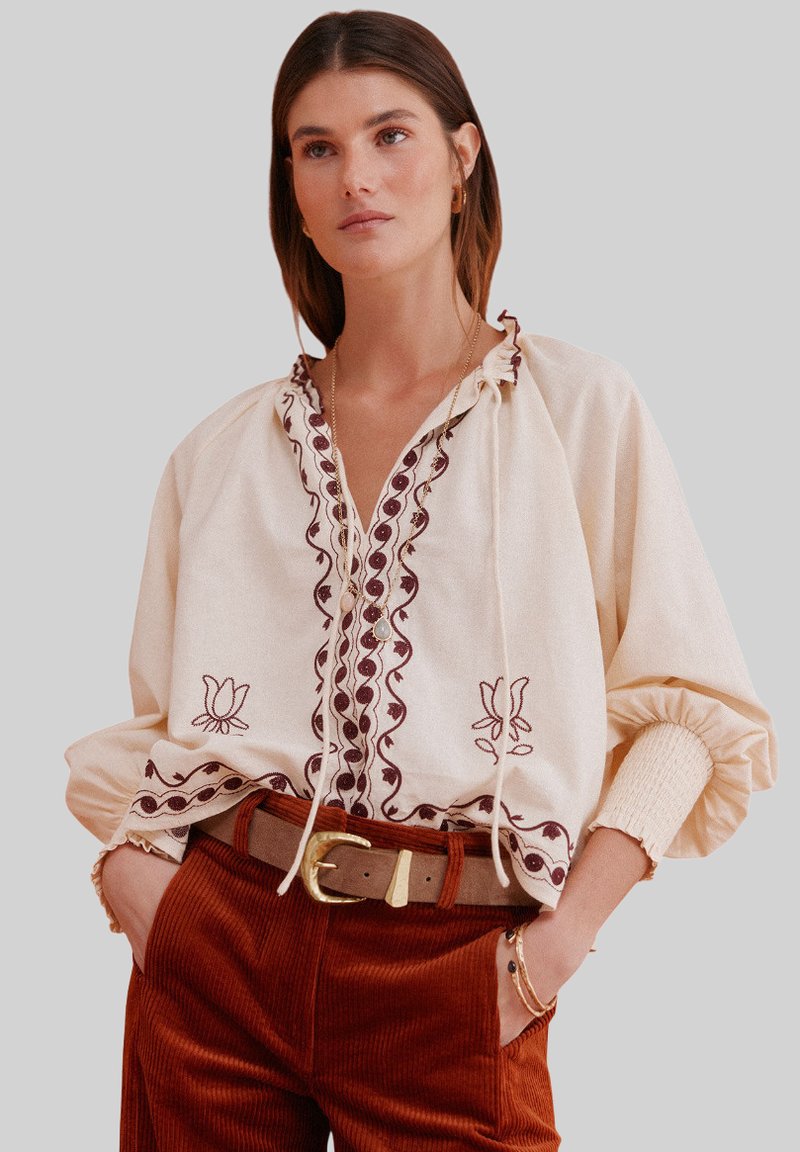 Cream blouse with embroidered detailing in burgundy, featuring a tie neck and puffed sleeves, worn with rust-colored corduroy pants and a belt.