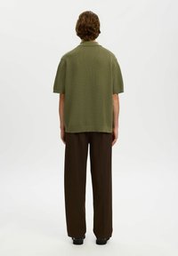 Personne aux cheveux bouclés bruns, vue de dos, portant un t-shirt tricoté à manches courtes vert olive et un pantalon ample marron foncé.