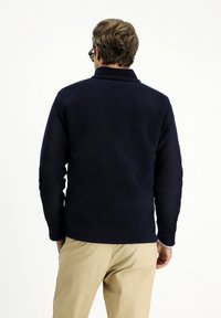 Granatowa polarowa kurtka z kołnierzem, długimi rękawami i gładką fakturą, w połączeniu z jasnobeżowymi spodniami. Widok z tyłu ukazuje prosty design.