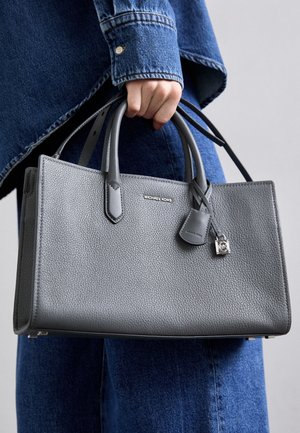 Ręka trzymająca szarą, teksturowaną torebkę Michael Kors z podwójnymi uchwytami i małym zawieszką w kształcie zamka, na tle niebieskich dżinsowych ubrań.