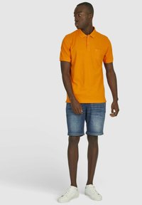 Oranje polo shirt met kraag en borstzak, gedragen met blauwe denim shorts en witte sneakers. Korte mouwen en effen kleur.