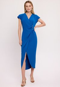 Niebieska sukienka w stylu kopertowym z krótkimi rękawami, z trzema dekoracyjnymi guzikami w pasie, rozcięciem po boku i gładką fakturą. Przezroczyste sandały na obcasie.