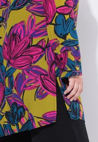 Květinová tunika s růžovými a modrými stylizovanými květinami na žlutém pozadí, s bočním rozparkem a knoflíkovými detaily na rukávu.