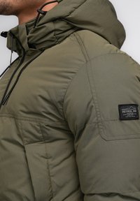 Jachetă puf verde măsliniu cu glugă, având un buzunar frontal și șnururi negre. Include o etichetă de marcă pe mânecă.