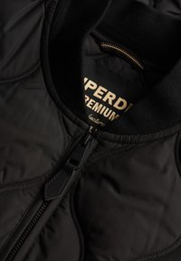 Zwarte gewatteerde jacket met een ribbelkraag, voorzien van een metalen ritssluiting en gouden branding op het binnenlabel. Zachte textuur.