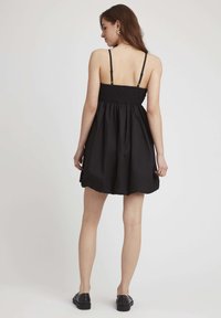 Robe noire avec un corsage froncé et des bretelles fines. Jupe évasée avec une texture unie. Portée avec des chaussures noires à enfiler.