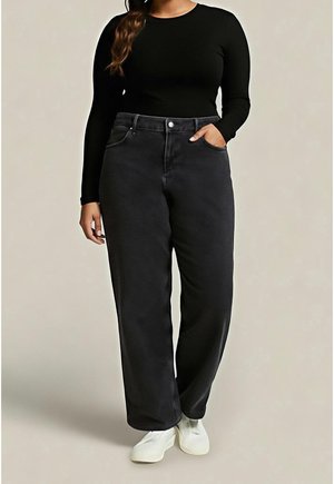 Femme portant un haut noir à manches longues, un pantalon noir ample avec une main dans la poche, et des chaussures blanches sans lacets, debout devant un fond uni.