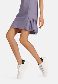 Sneakers bianchi con accenti neri sul tallone, dotati di occhielli e lacci argentati. Indossati con un vestito lavanda con orlo arricciato.